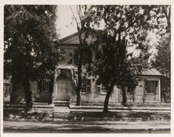 View of a house in a residential area, obscured by trees. "The house was built in 1835 by Ellis and was last occupied by the Misses Bessie and Barbara Hagemeister. It was located on the northeast corner of Pine and Adam Streets where the Northland Hotel now stands."