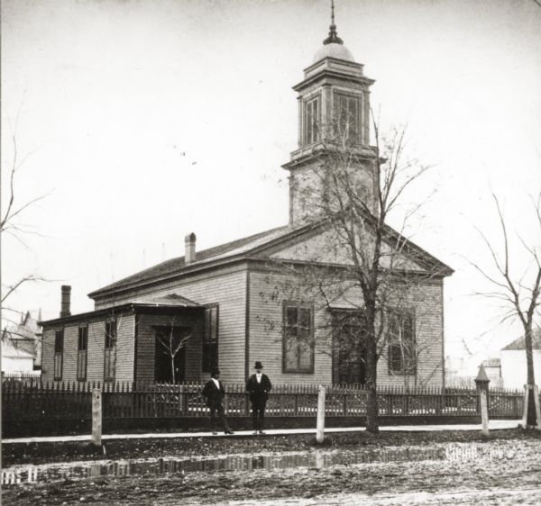 View from unpaved street towards a church. Two men are standing ont he sidewalk in front of the picket fence in front of the church. The church was built in 1838, but was destroyed by fire about 1880.