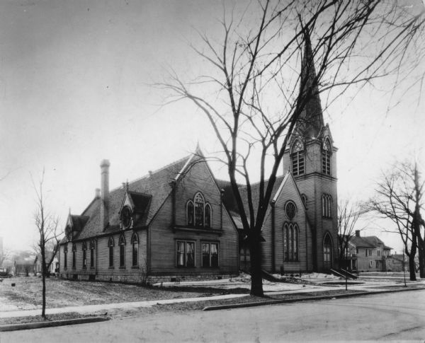 View of the church. A dusting of snow is on the church lawn. The church has a tall spire, and stained-glass windows with gothic arches. First Presbyterian Church (later Union Congregational), the second church building, seen after remodeling.