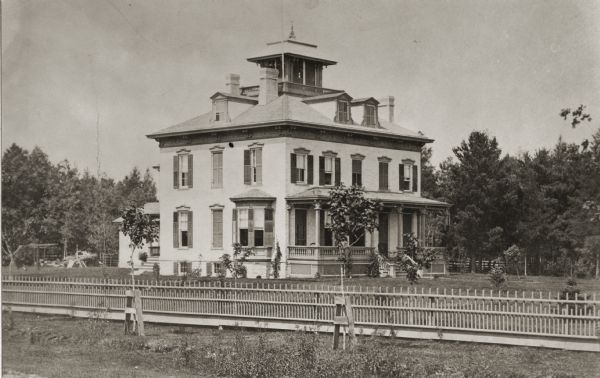 Exterior of home and lawn on a summer day, with a forest in the background and a picket fence in the foreground. The house has a lookout on the roof.