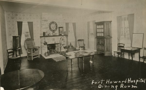 View of a room with hardwood floors, with a wooden, four-seater table in the center of the room. A wooden, oval-shaped table is in the left foreground. Five chairs and a desk are in the room, as well as a wooden cabinet. A fireplace is on the back wall with a circular-shaped portrait hanging over it.