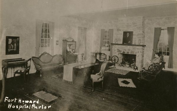 View of a parlor built partially out of stone. A fireplace is on the back right wall, with a portrait hanging above it, and a spinning wheel near the left side of it in the corner of the room. A woman is sitting in a chair on the right near the fireplace. A wooden table stands in the center with a cloth draped over it. Four rugs are on the floor, and a small piano is against the left wall.