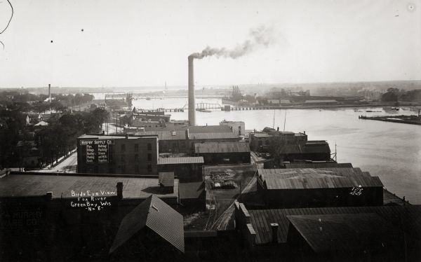 Elevated view over rooftops towards the Fox River looking south, with factories and a smokestack in the foreground.