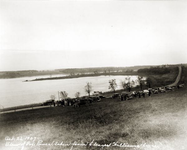View down hill towards the Fox River from the Eleazer Williams home. A group of people are automobiles are near a road near the shoreline.