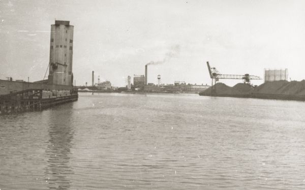 View of the harbor. On the left is a grain elevator and wood pulp on the wharf.  In the center background is the Northern Paper Mills and to the right are the coal yards for up-river traffic.