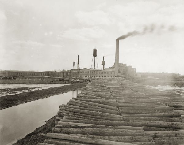 View of the Green Bay (later Hoberg) Paper and Fibre Company, from the back. Stacks of logs are in the foreground near water.