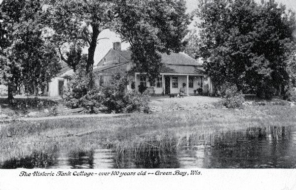 View across water toward the Tank Cottage. Caption reads: "The Historic Tank Cottage — over 100 years old — Green Bay, Wis." 