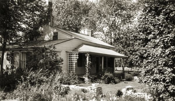View of the cottage from the outside, with trees and plants surrounding it. A garden is in the foreground with a low-lying brick wall. The cottage's porch has lattice on it. On the original site on the west side of the Fox River before restoration.