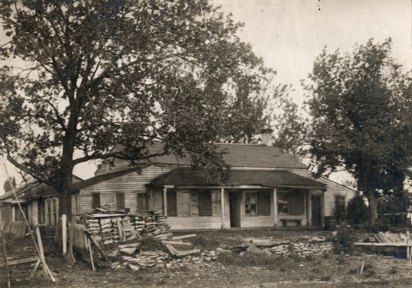 A view of the cottage with large trees around it, and a stone wall in the front yard. Some windows appear shattered. Wooden pallets are stacked in the front yard, along with other debris. A woman in a dress is barely visible on the far right. This is the original site on the west side of the Fox River, before restoration.