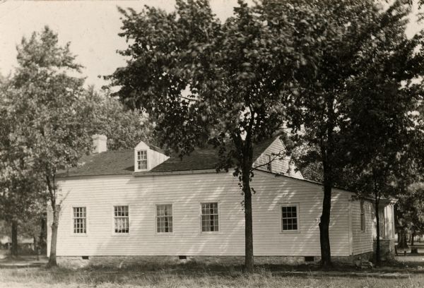 View of the broadside of the cottage during warm season, with a few large deciduous trees casting shadows across the cottage, though it's a sunny day. On the original site on the west side of the Fox River before restoration.