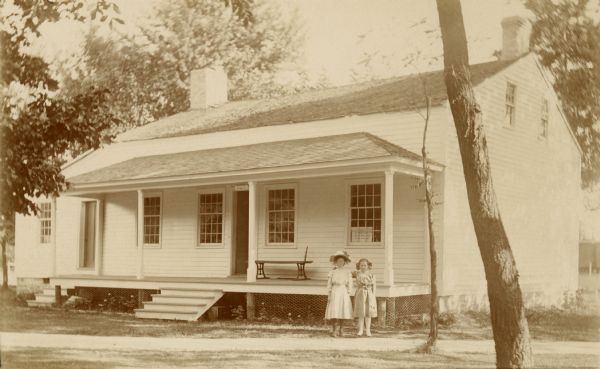 Exterior view of the cottage and porch. Two little girls in dresses stand in front of the cottage, posing for the camera. A bench stands on the porch. On the original site on the west side of the Fox River, before restoration.