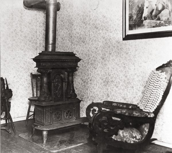 Interior of the cottage with a rocking chair, a wood stove, a painting on the wall, and a spinning wheel on the left. The floor is wooden, and the wallpaper has a floral pattern.