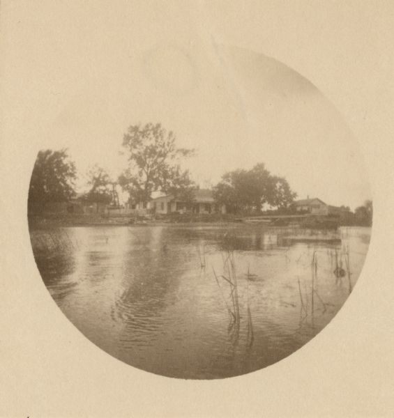 Distant view of the cottage across the river, with reeds in the foreground. On the original site on the west side of the Fox River, before the restoration.