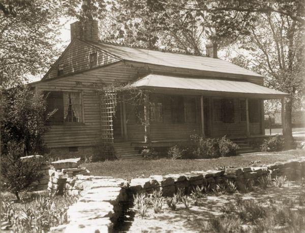 View of the cottage with a stone wall in front of it, and surrounded by plants and trees. On the original site on the west side of the Fox River before restoration.