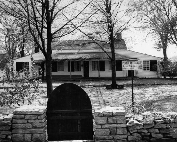 Front view of the Tank Cottage with a stone fence and gate in the foreground. The cottage was built in 1776 by Joseph Roi, a fur trader, who lived in it until 1805 when it was purchased by Jacques Porlier. Later, the Tank family, who presented Tank Park to the city, lived in it. In recent years, the Brown County Historical Society and the South Side Improvement Association have rescued it from an industrial section of the city and taken it to Tank Park, where it now stands