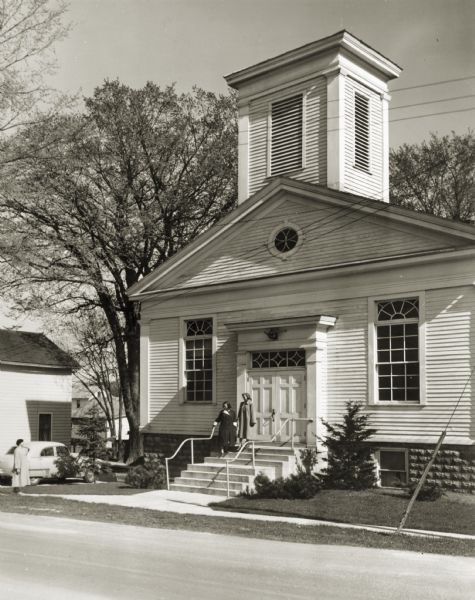 View of the First Methodist Church, with two women standing on the church steps, and another woman standing on the sidewalk. The church was built in 1854 by Charles Robinson, the builder of Butternut House (located across the street from the church) and possibly the designer of the Old Wade House.