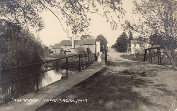 View of a bridge with several buildings in the background. Caption reads: "'The Bridge' Honey Creek, Wis."