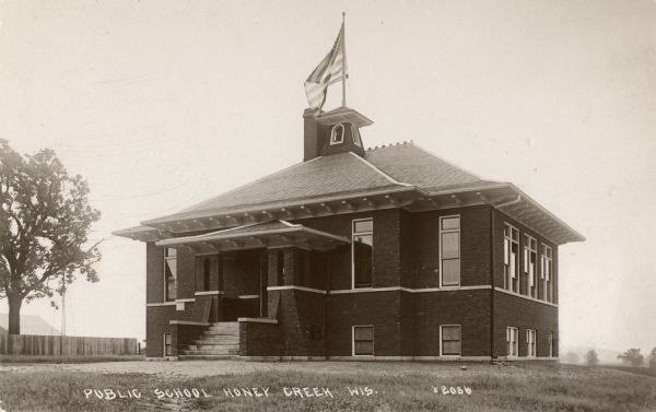View towards front and right side of a public school. A U.S. Flag is flying from the top of the belltower. Caption reads: "Public School Honey Creek Wis."