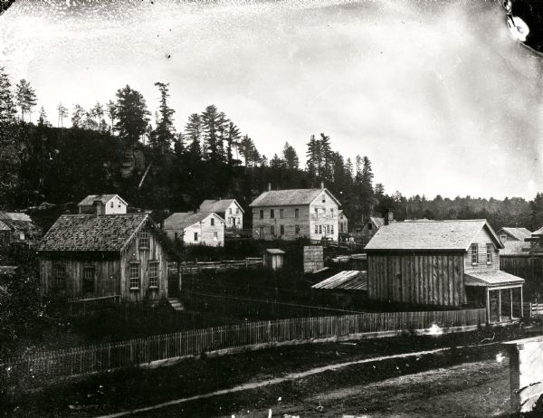 An ambrotype of Wonewoc. A road and fence is in the foreground, with houses beyond. A tree-lined hill is in the background.
