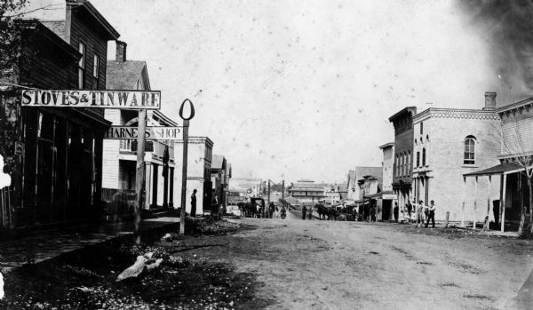View down unpaved street in Winneconne towards the bridge from the east side of town. Storefronts on the left have signs for "Stoves & Tinware" and "Harness Shop".