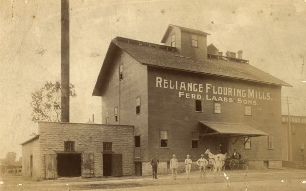 The Reliance Flooring Mill. Men are posing in front, and another man is on a horse-drawn vehicle near the loading dock. The mill building was physically moved by water by Capt. Sam Neff from Winnconne to Oshkosh, Wis. July 4, 1879.