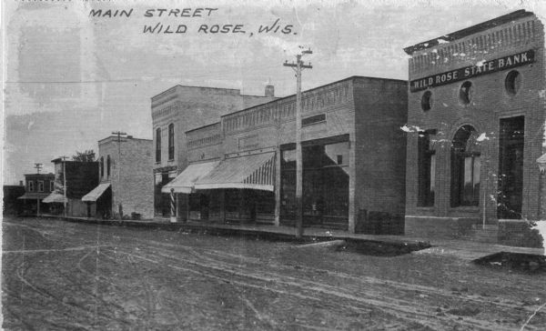 View across unpaved street towards commercial buildings and storefronts on the right. The Wild Rose State Bank is on the far right. Caption reads: "Main Street Wild Rose, Wis."