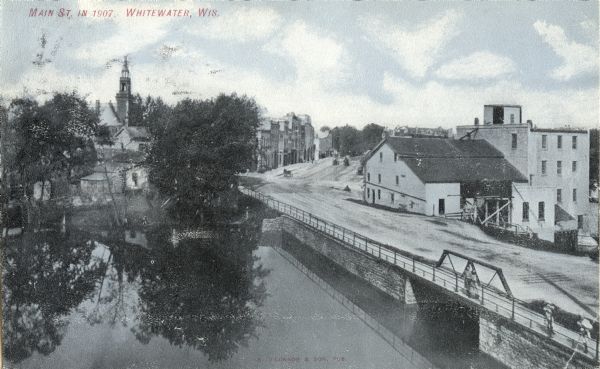 Elevated view over water towards Main Street with bridge. Caption reads: "Main St. in 1907, Whitewater, Wis."