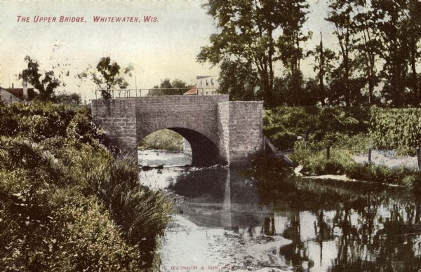 View from bank towards the stone bridge. Caption reads: "The Upper Bridge, Whitewater, Wis."