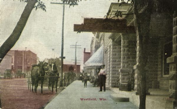 A view down the sidewalk with storefronts on the right. Pedestrians are standing on the sidewalk, and horse-drawn vehicles are along the curb. Caption reads: "Westfield, Wis."
