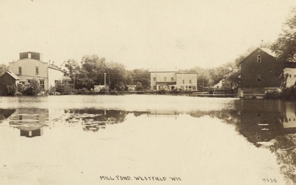 Looking across Mill Pond towards buildings along the shoreline. Caption reads: "Mill Pond, Westfield, Wis."