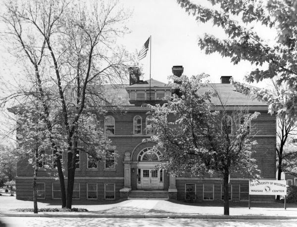 Front view of the University of Wisconsin, Wausau Center, with a flag atop the building and trees in the foreground.
