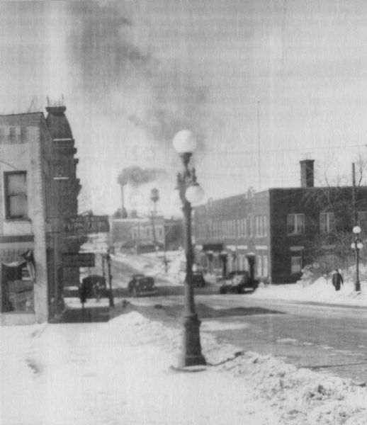 Winter view of a city street with parked automobiles and people walking down the sidewalk. A lamppost is in the foreground, and a smokestack and water tower are in the background. Snow is on the ground.
