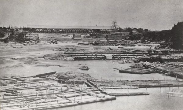 Elevated view taken from the extreme end of Clark's Saw and Planing Mill. An old flour mill can be seen behind the bridge, and to the extreme right are three small stores. One is a tobacco store owned by Joe Habeck. There are rock footings for three piers of the C. & N.W. R.R. bridge. In the foreground are lumber rafts whose destination eventually will be St. Louis, Mo.