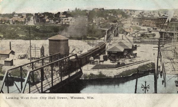 Elevated view of the railroad station looking west. A bridge over the river is in the foreground, and a large water tower is across the railroad tracks on the left. Caption reads: "Looking West from City Hall Tower, Wausau, Wis."