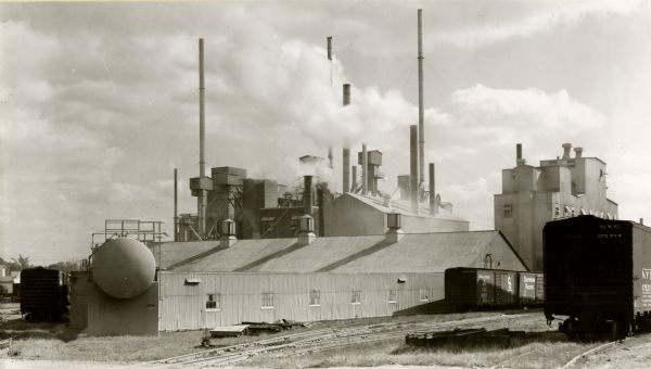 View of the Minnesota Mining and Manufacturing Company plant. The sky is cloudy and smoke obscures some of the stacks. Railroad cars can be are on the right.