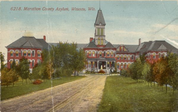 View looking down the dirt road framed by trees toward the Marathon County Asylum. Caption reads: "Marathon County Asylum, Wausau, Wis."