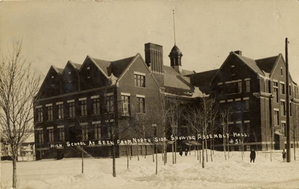 View of the high school from the north side showing the Assembly Hall.  Snow is on the ground, and a few people are standing among the small trees in front. Caption reads: "High School as seen from North side showing Assembly Hall."