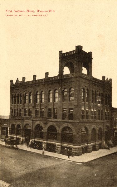 Elevated view of the First National Bank. There are people standing on the pavement outside the bank and a horse-drawn carriage is parked on the street.
