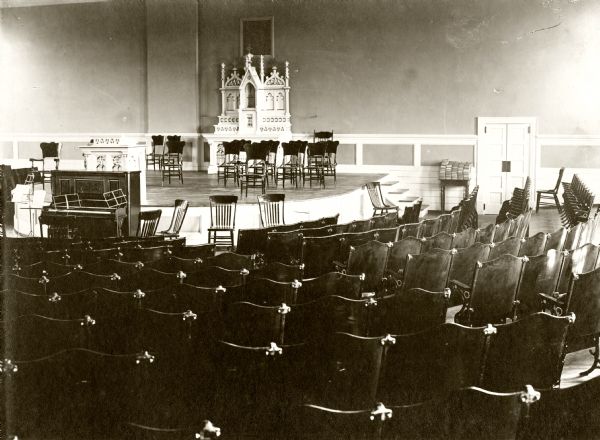 View of the chapel in the Wisconsin State Prison. In the foreground are chairs, and in front of the stage is a piano.