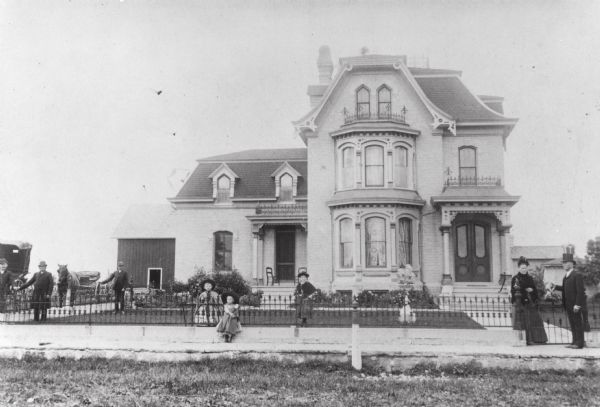 View of the M.K. Dahl residence with several children and adults standing in front of it along a fence.