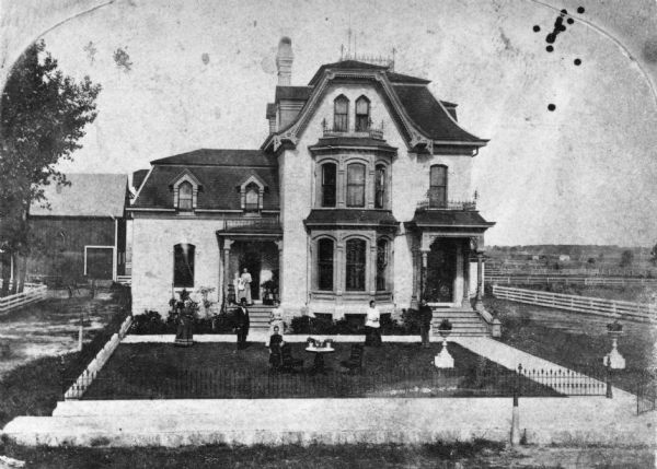 Slightly elevated view of the M.K. Dahl residence. People are posing in the yard. There is a barn in the background on the left.
