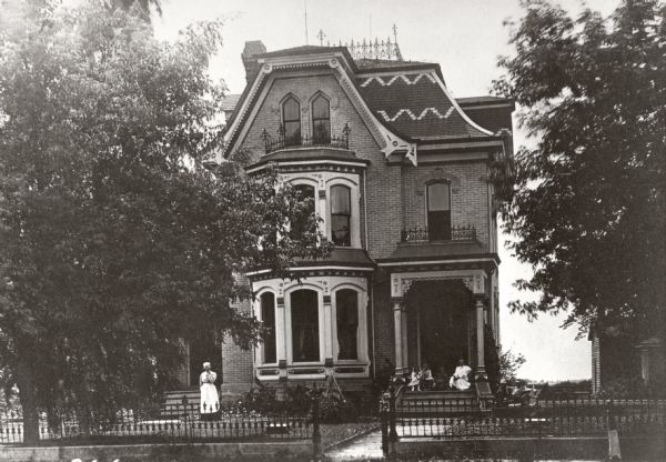 View from street towards the M.K. Dahl residence, with several children seated on the front porch, and a woman standing on the left.