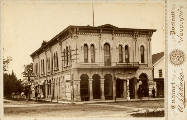 The Opera House at the corner of Washington (now Main) and North Mill Streets. A sign at the back of the building reads: "Printing." A barber pole is in front. Pedestrians are on the sidewalk.