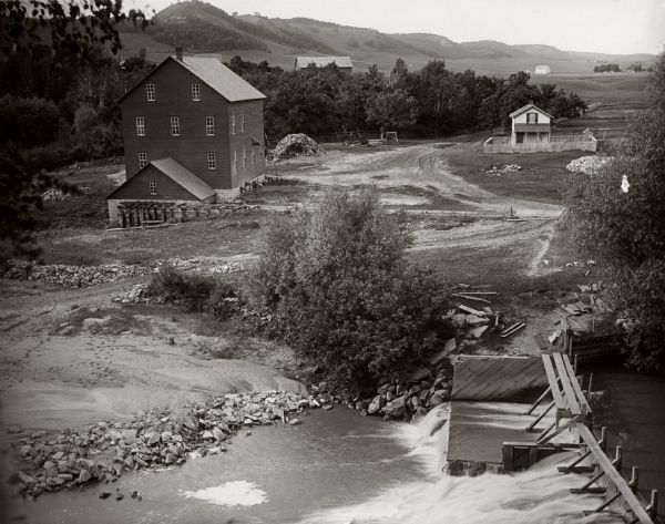 View of a mill, sluice and dam on Waumandee Creek. The mill was built by John Oschner in 1863. Two products produced by the mill were White Rose Flour and Wheat Gitz Cereal. A home is visible in the distance.