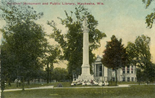 View of the Soldiers' Monument in front of the Waukesha Public Library, in the center of Cutler Park. A group of people are sitting on the lawn in front of the monument. Caption reads: "Soldiers' Monument and Public Library, Waukesha, Wis."