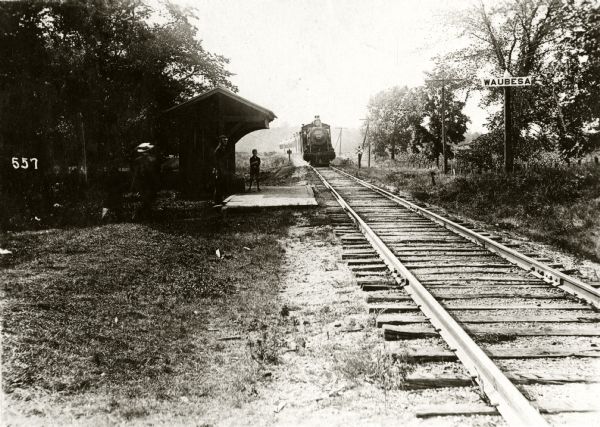 View down left side of railroad tracks towards a railroad station with several figures standing on and around the platform. A sign on the right reads: "Waubesa."