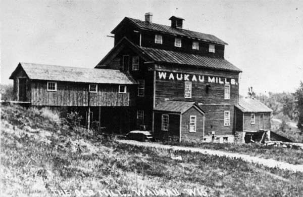 Exterior view of the Bean and Palfrey flour mill. The sign on the building reads: "Waukau Mills". Caption reads: "The Old Mill, Waukau, Wis."