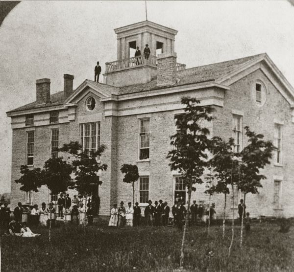 View of Carroll College, with groups of men and women standing outside of the building.
