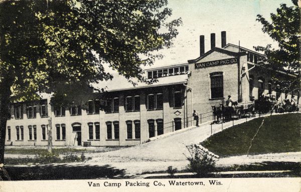 View of the Van Camp Packing Company. People, including a horse-drawn vehicle are on a driveway on a hill near the side of the factory. Caption reads: "Van Camp Packing Co., Watertown, Wis."