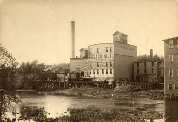 View across water towards the Empire Mill. Alongside the building are railroad cars at a loading dock. The signs on the mill read: "Globe", "Globe's Empire" and "Custom Mill".